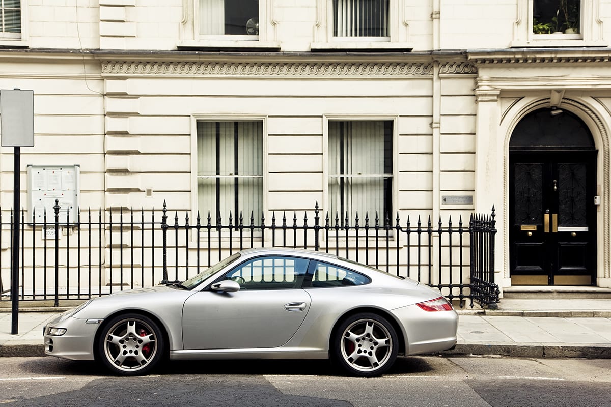 Porsche steht auf einer dreckigen Straße vor einem Metallzaun und einem beigen Haus | vor der Retusche